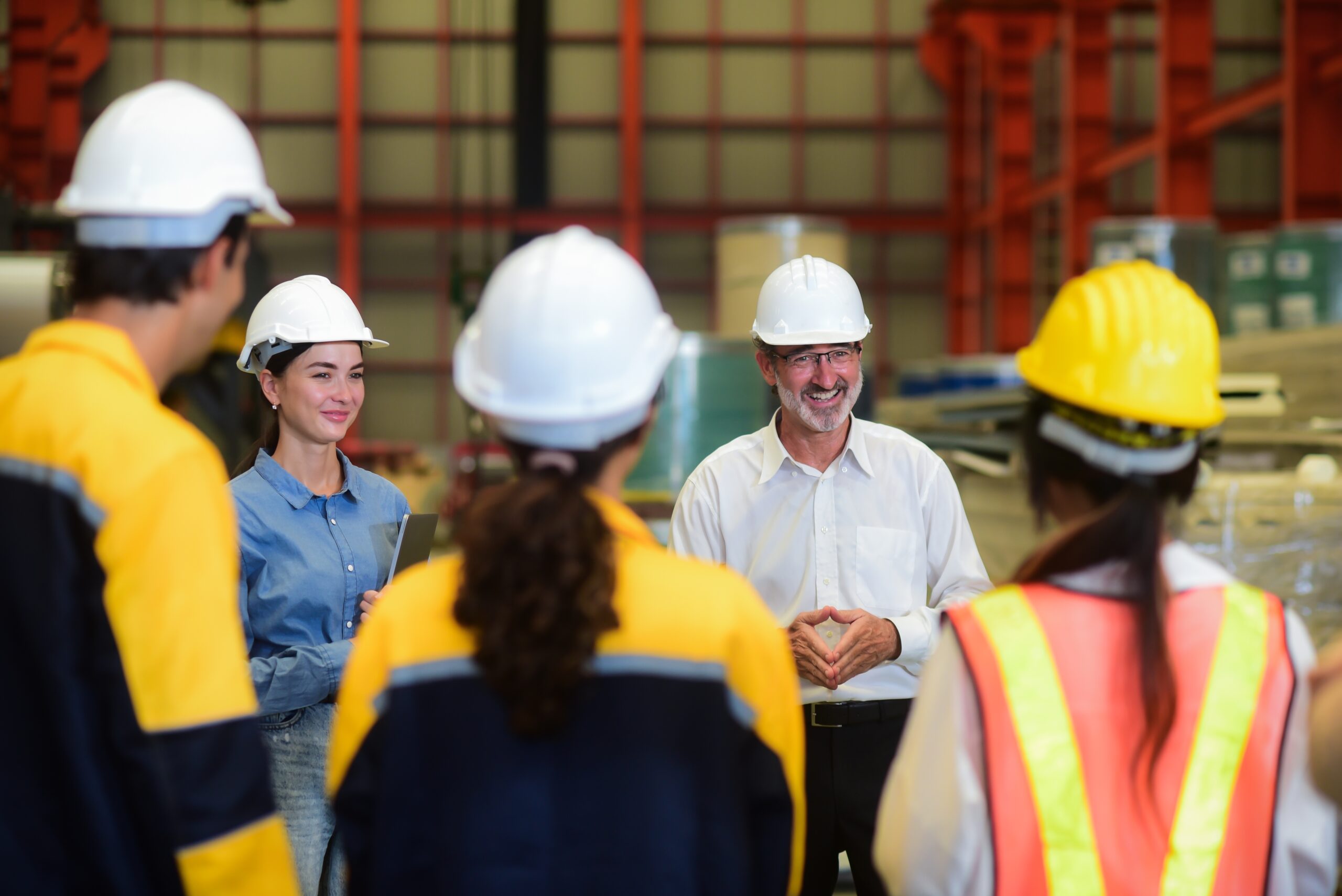 Safety manager leads a team meeting with industrial workers in hard hats and high-visibility vests inside a manufacturing facility, discussing onsite OSHA and MSHA compliance procedures. Safety manager leads a team meeting with industrial workers in hard hats and high-visibility vests inside a manufacturing facility, discussing onsite OSHA or MSHA compliance procedures.